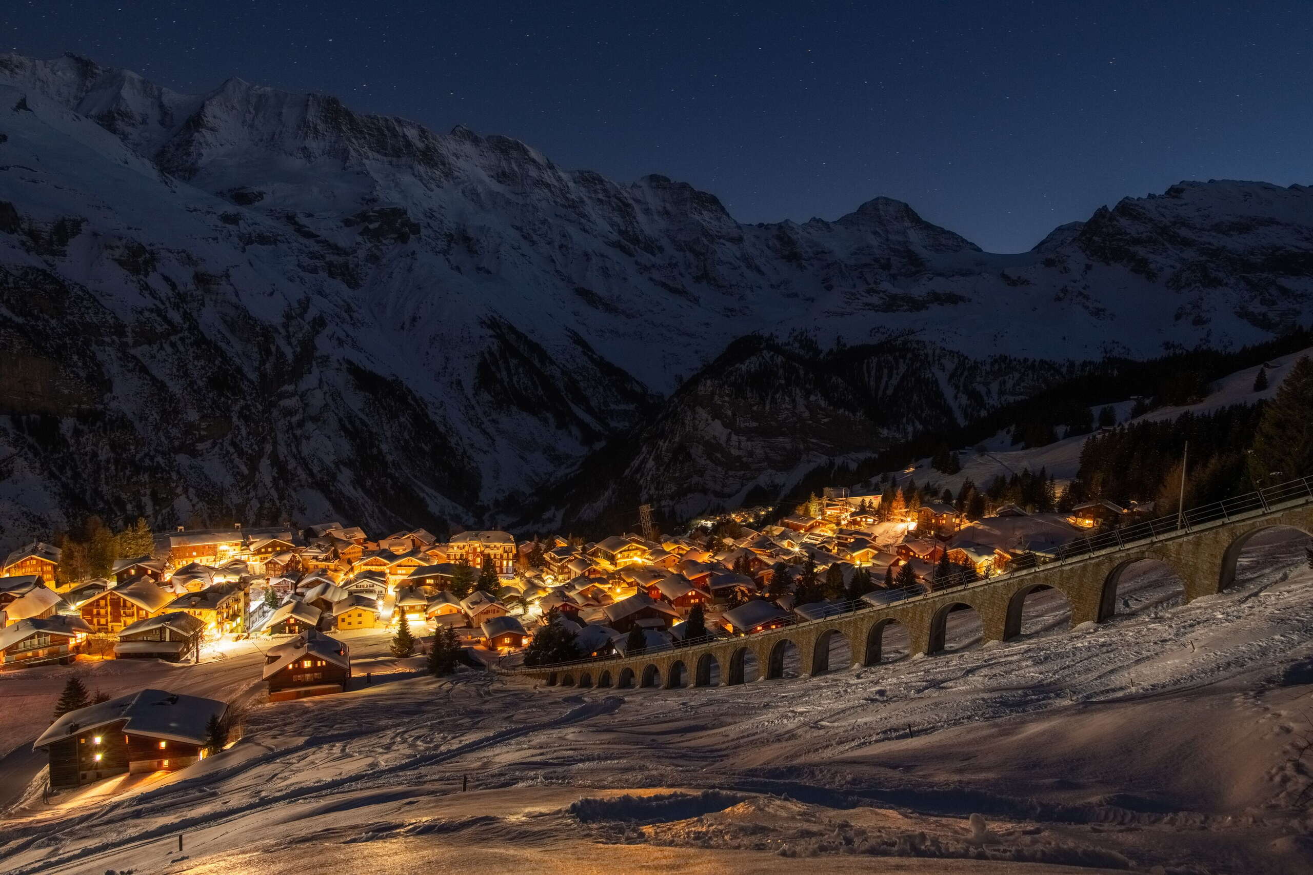 La nuit, le village enneigé de Mürren s'illumine d'une couleur dorée. En arrière-plan, on distingue à peine le panorama des montagnes dans l'obscurité.