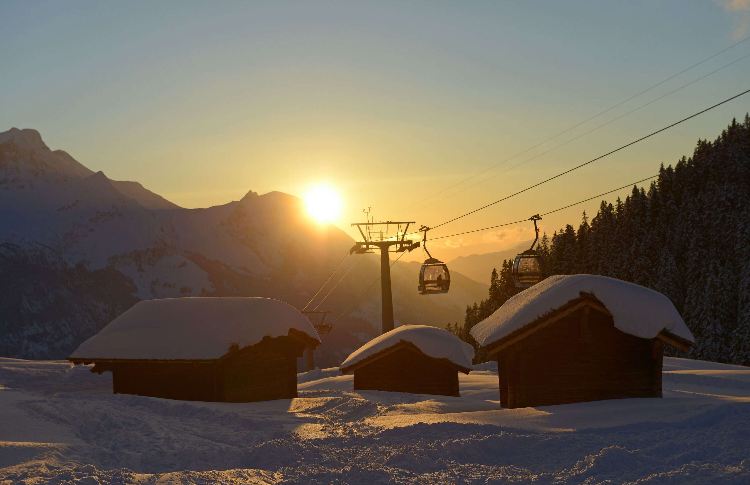 Two gondolas of the Meiringen-Hasliberg mountain railroads at sunset, with three snow-covered huts in the foreground. Taken at the Bidmi intermediate station.