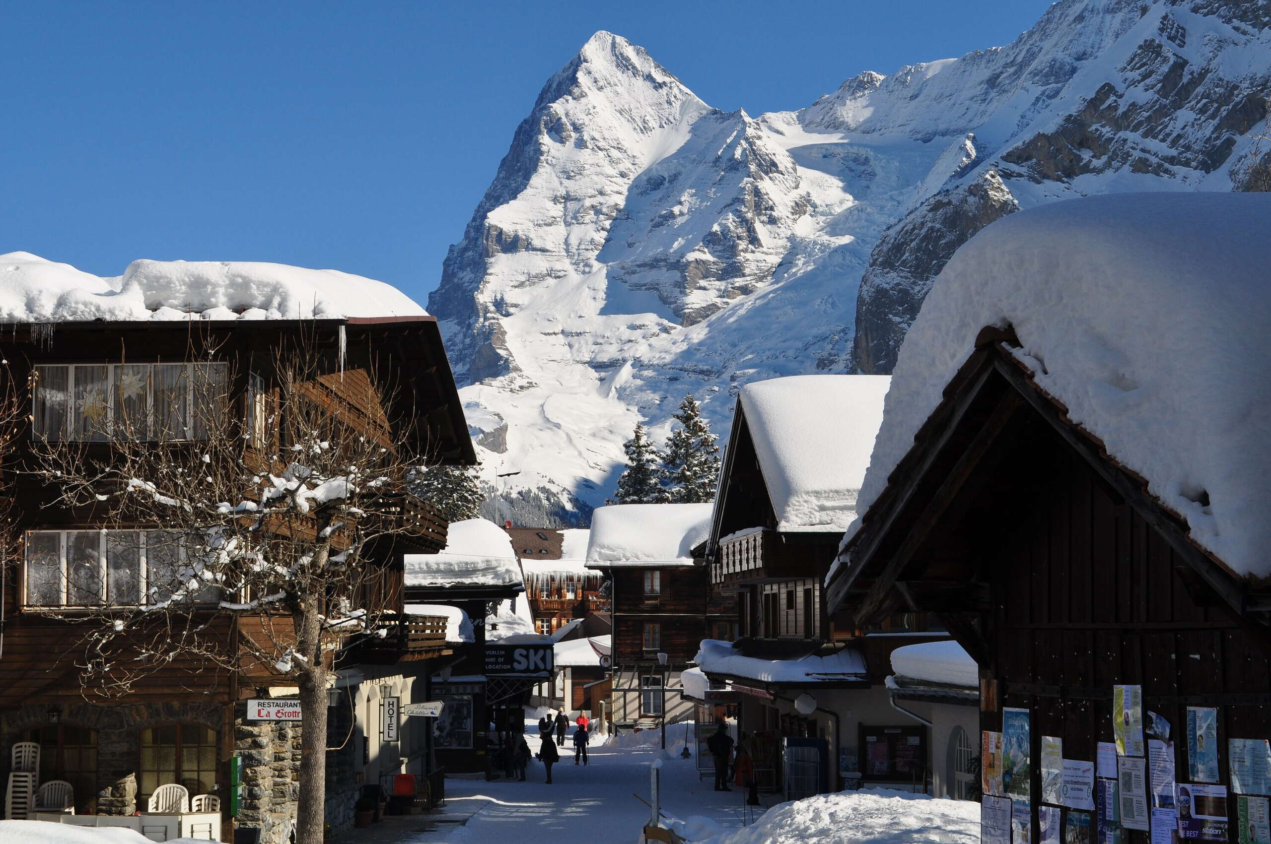 Personen flanieren durch die schneebedeckte Dorfstrasse. Der schneebedeckte Eiger ragt im Hintergrund empor.