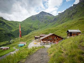 Wide-angle shot of the Schiltalp mountain lodge and alpine cheese dairy, with a flagpole on the left. The Schilthorn is visible in the background.