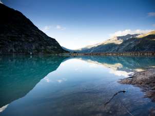 Le lac de Gelmer est niché au cœur du paysage de haute montagne du massif du Grimsel. La surface calme de l'eau reflète les flancs des montagnes environnantes et le ciel limpide. Au premier plan, on aperçoit les rochers des rives, tandis que le barrage délimite le lac à l'arrière-plan.