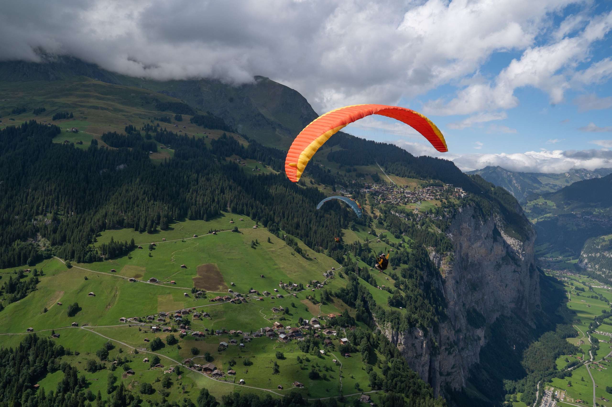 Mehrere Gleitschirme fliegen über grüne Matten und das Tal mit verstreuten Häusern.